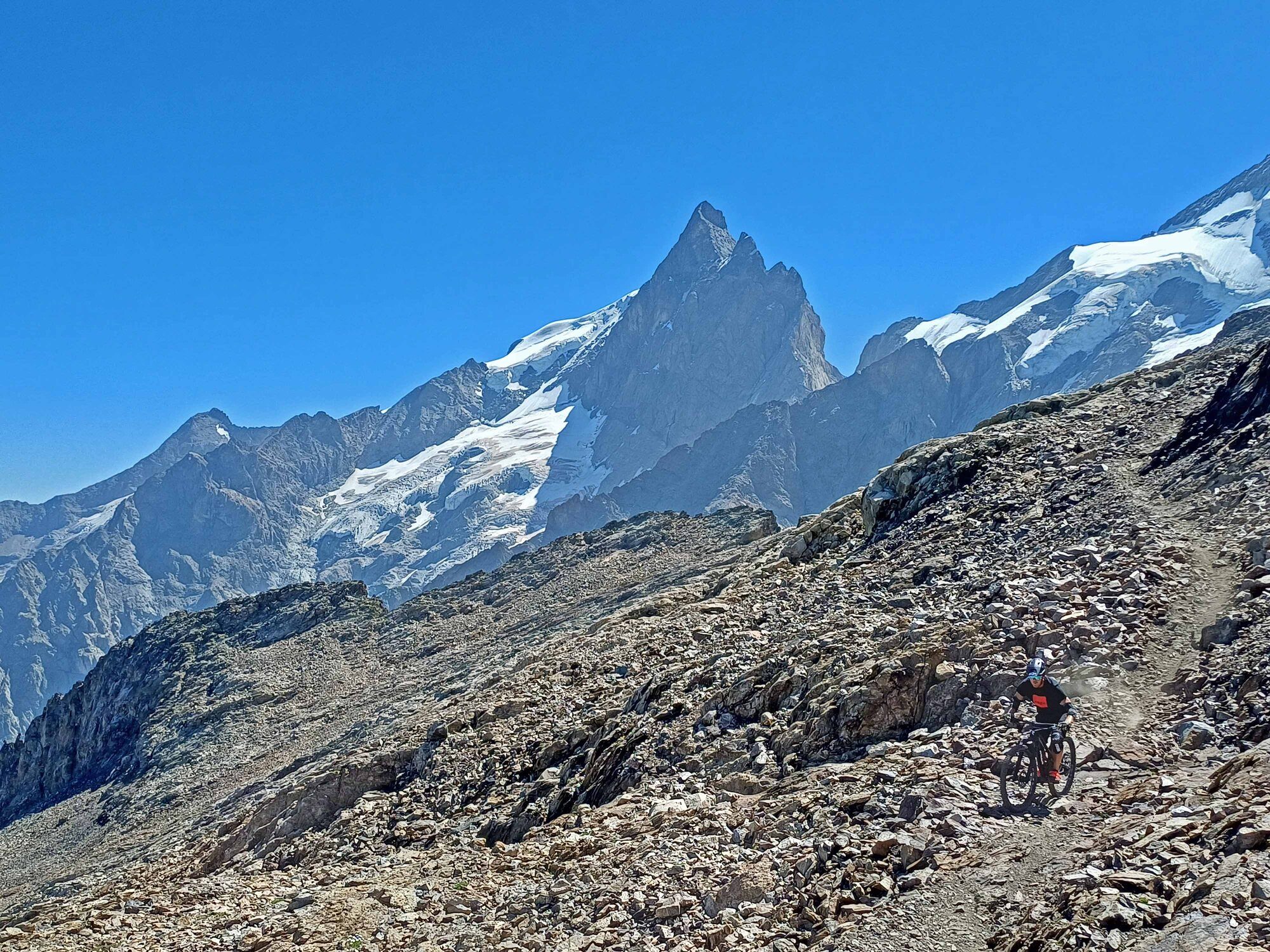 Mountain biker riding a rough rocky trail beneath a snow-covered Alpine peak