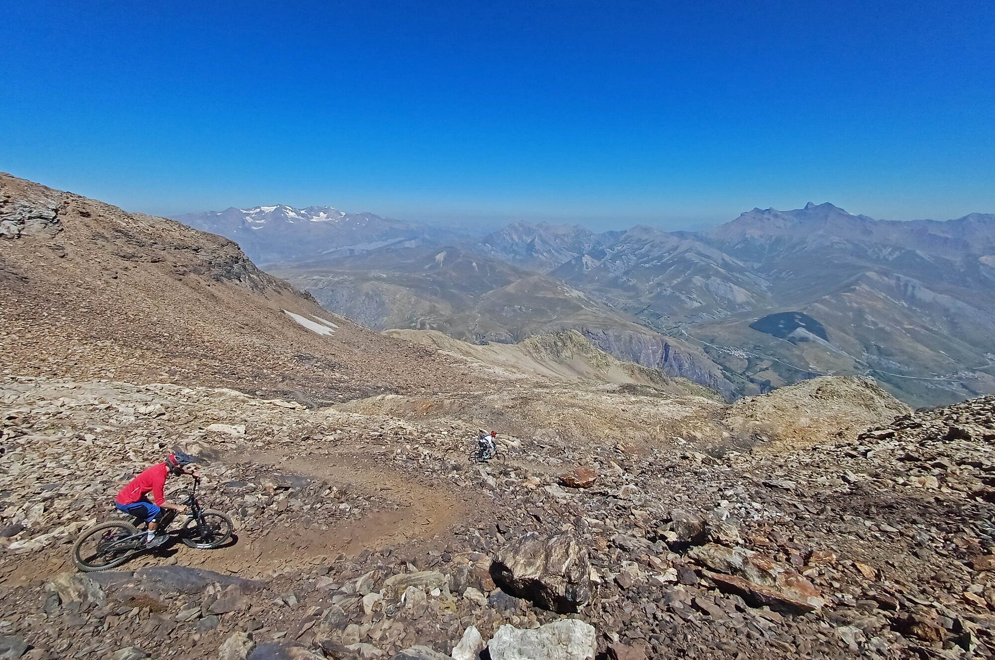 Two mountain bikers descending a rocky high-Alpine trail above a wide mountain valley