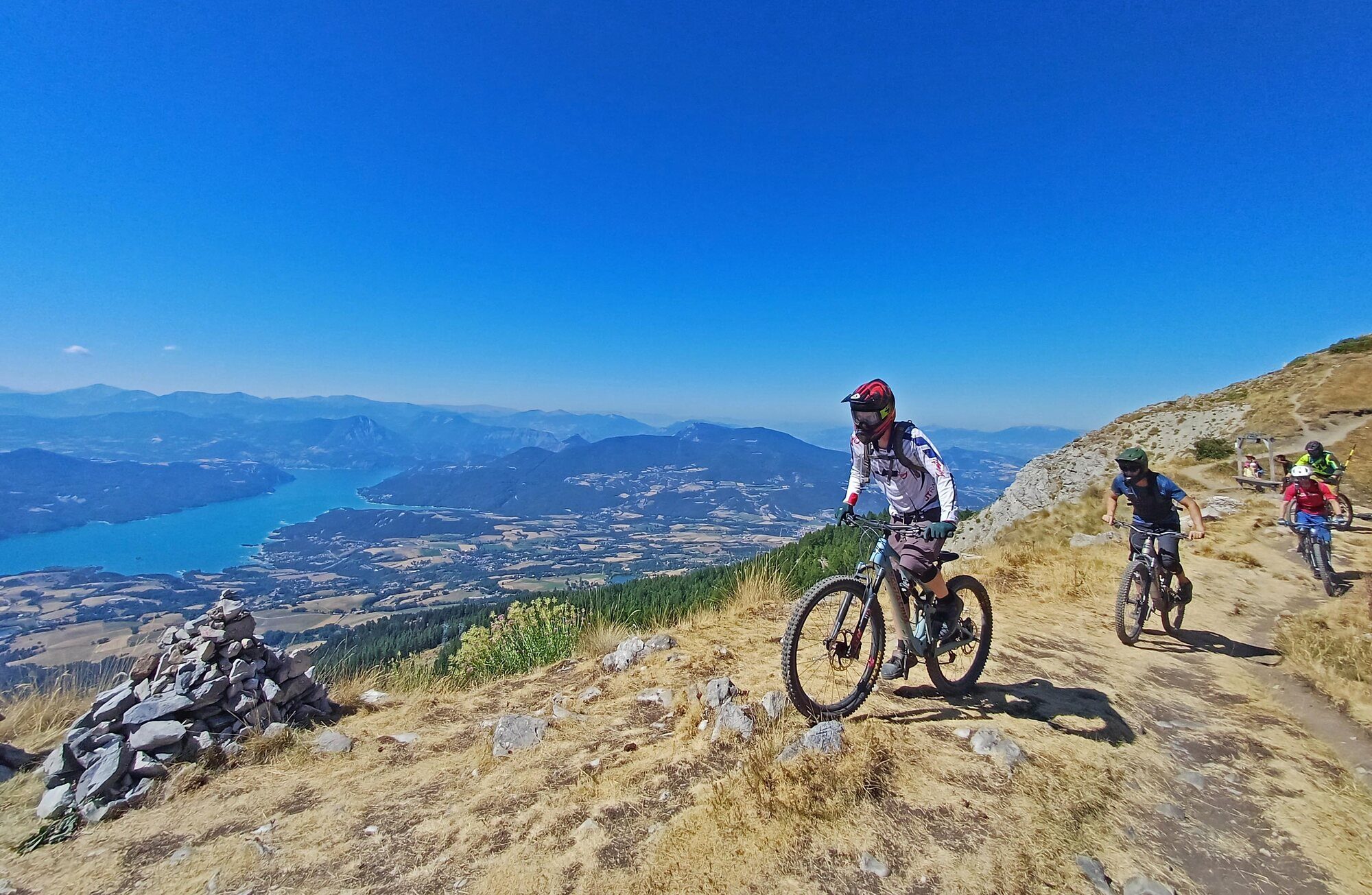 Mountain bikers traversing a sunny high-Alpine trail above a lake and broad mountain valley