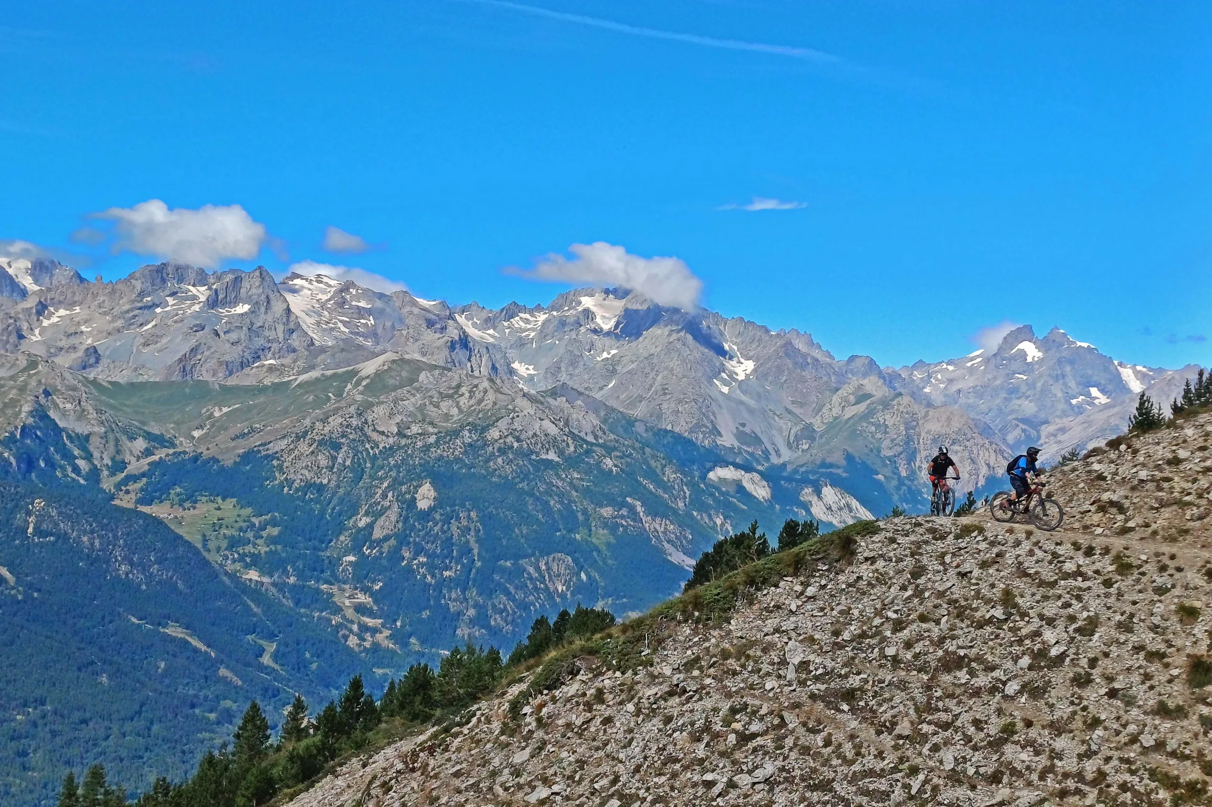 Two riders on a rocky high Alpine trail with snowy peaks in the distance