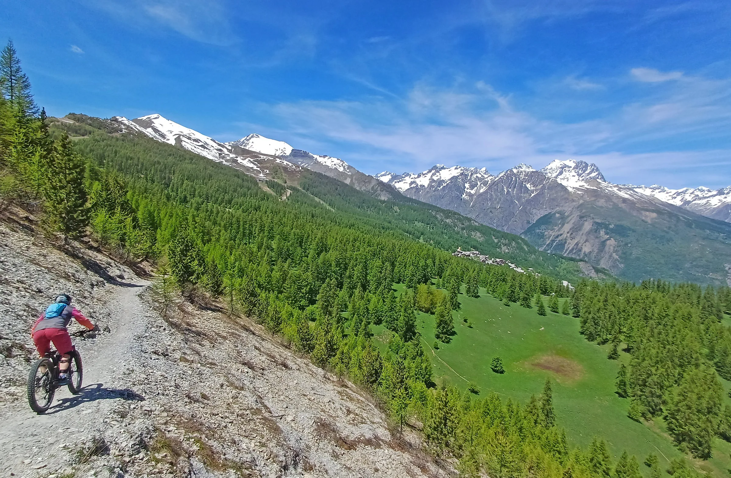 Rider descending a rocky trail below snow-capped peaks and green forests